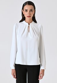 Blusa bianca a maniche lunghe con dettagli plissettati sulle spalle, scollatura a chiave e un bottone decorativo al collo, abbinata a pantaloni neri.