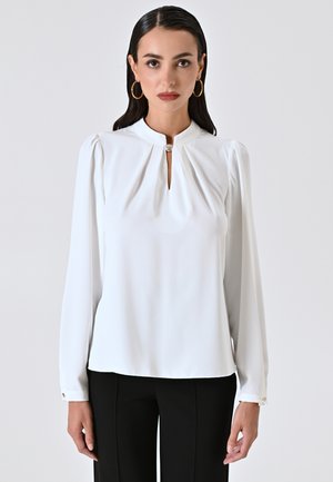 Blusa bianca a maniche lunghe con dettagli plissettati sulle spalle, scollatura a chiave e un bottone decorativo al collo, abbinata a pantaloni neri.