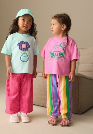 Two young children stand indoors; one wears a mint hat and flower shirt with pink pants, the other wears a pink "Home is my happy place" shirt and striped pants.