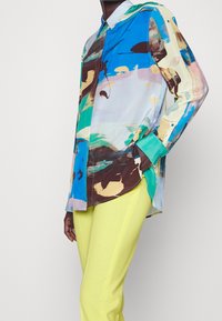 Flerfärgad långärmad skjorta med abstrakta mönster i blått, grönt, brunt och krämfärgat. Inkluderar en knapp-up krage och manschetter. Kombinerad med ljusgula byxor.