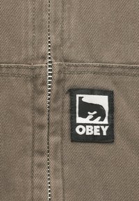 Siva tkanina s finom dijagonalnom teksturom, koja ima vertikalni patentni zatvarač i crnu pravokutnu etiketu s bijelim grafičkim dizajnom i tekstom "OBEY".