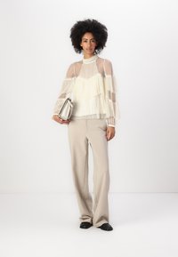 Sheer cream top with layered ruffles, long sleeves, and a high neck; paired with beige wide-leg trousers and black loafers.