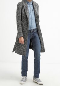 Abrigo largo de color gris jaspeado con abertura frontal, llevado sobre una camisa de mezclilla de color azul claro, combinado con jeans azules y zapatillas plateadas. Textura suave.