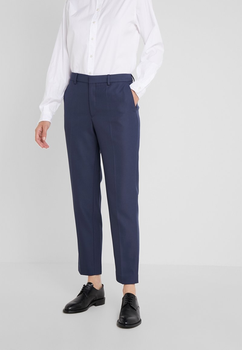 Personne portant un pantalon ajusté bleu marine, une chemise blanche boutonnée et des chaussures habillées en cuir noir, debout avec une main dans la poche.