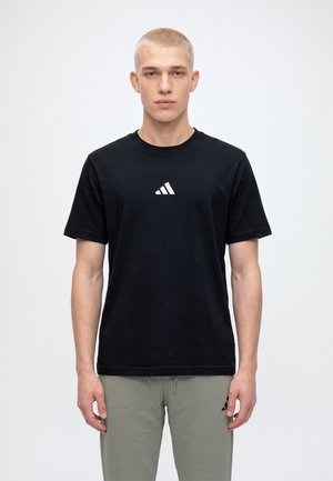 Мъжки модел стои с лице напред, облечен в черна тениска Adidas и сив панталон, на изчистен светъл фон.
