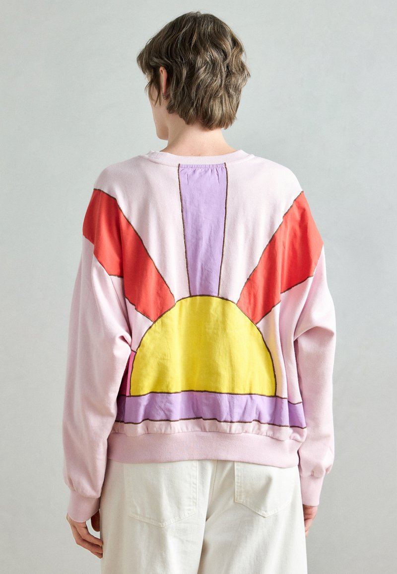 Ružová mikina s grafickým motívom žltého slnka s viacfarebnými lúčmi v červenej a fialovej farbe. Mäkká, štruktúrovaná látka s pohodlným strihom.