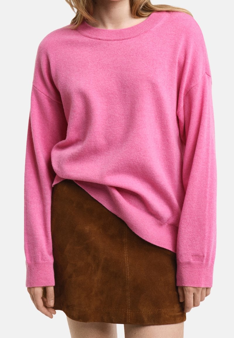 Femme portant un pull ample rose à manches longues rentré dans une mini-jupe en daim marron, sur un fond uni.