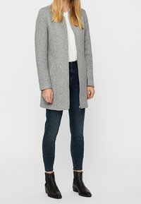 Harmaa villasekoitetakki suoran siluetin, pitkien hihojen ja siistin kaula-aukon kanssa, yhdistettynä tummiin farkkuihin ja mustiin nilkkureihin.