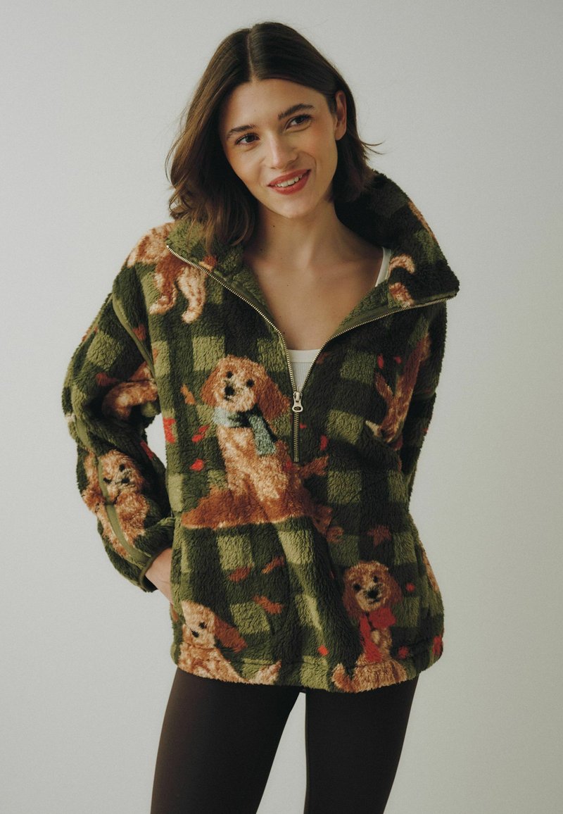 Woman wearing green fleece pullover with dog patterns standing against plain light background, smiling slightly with hands in pockets.