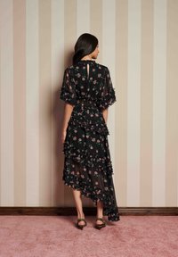 Kvinna i svart blommig klänning med volanger, stående barfota på rosa matta mot en beige randig vägg, vänd bort från kameran.