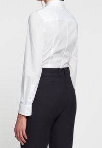 Camicia bianca aderente con colletto alto, polsini con bottone e pieghe sul retro, abbinata a pantaloni scuri su misura con vita con cintura.