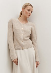 Licht beige gebreide cardigan met lange mouwen, ronde halslijn en zes goudkleurige knopen aan de voorkant. Zachte, gestructureerde stof.