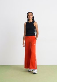 Svart ermeløs topp, løse røde bukser med hvite side-striper, og grå joggesko. Glatt stoff, minimalistisk design, og kontrasterende farger.