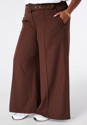Femme portant un pantalon marron taille haute, jambe large, avec ceinture et baskets blanches, main dans la poche, sur fond blanc uni.