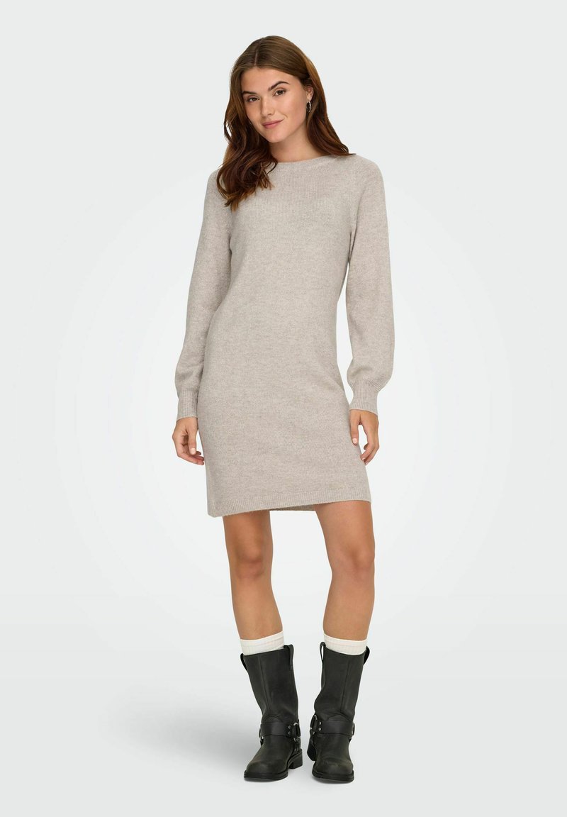 Femme debout portant une robe en tricot beige à manches longues, des bottes noires mi-mollet et des chaussettes blanches sur un fond clair neutre.