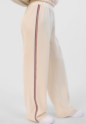 Kremowe spodnie o szerokich nogawkach z bocznymi czerwono-biało-czarnymi paskami, noszone z białymi trampkami.