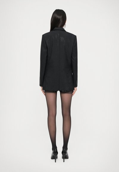 Blazer nero con paillettes, dal taglio sartoriale, maniche lunghe e rever a punta. Indossato sopra una mini gonna nera, abbinato a collant neri trasparenti e tacchi alti.