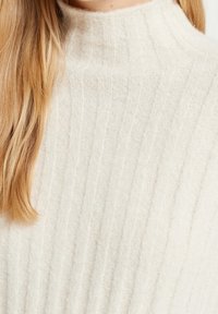 Pull en maille couleur crème avec col montant, texture côtelée et tissu doux et léger. Petites taches subtiles visibles dans le matériau.