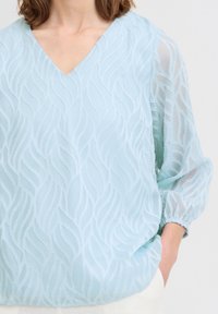 Blusa blu chiaro con scollo a V in tessuto trasparente e texture con motivi a onda. Maniche lunghe a sbuffo con polsini elasticizzati.