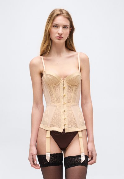 Γυναίκα που φορά μπεζ κορσέ από δαντέλα με μπροστινά κλείσιμο, ταιριαστά μπεζ τιράντες ζαρτιέρας, μαύρες δαντελένιες κάλτσες μέχρι το μηρό και καφέ εσώρουχο.