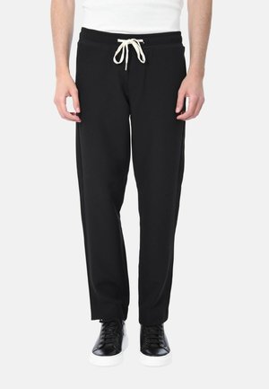 Uomo che indossa pantaloni neri con coulisse e sneakers nere con suola bianca, in piedi contro uno sfondo bianco uniforme.