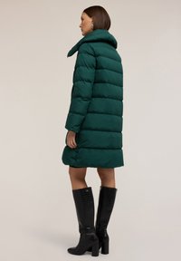 Zimní bunda v tmavě zelené barvě, s prošívaným vzorem, vysokým límcem a dlouhými rukávy. Nosit s černými vysokými botami na kolena a hladkým povrchem.