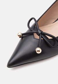 Chaussure en cuir noire pointue avec un nœud attaché comportant deux accents de boules dorées brillantes sur fond blanc.