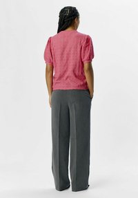 Blouse rose en dentelle avec des manches bouffantes, associée à un pantalon large gris. La blouse présente un motif texturé, tandis que le pantalon a une coupe ajustée.
