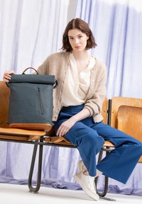 Rolltop-Rucksack aus dunkelblauem Material mit Reißverschluss und braunen Lederakzenten. Modell sitzt auf einem Holzstuhl, trägt eine beige Strickjacke und blaue Hose.