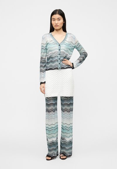 Cardigan z dolgimi rokavi in gumbi v vzorcu cik-cak, ki vključuje odtenke modre, črne in bele barve, kombiniran s hlačami širokih hlačnic v istem vzorcu.