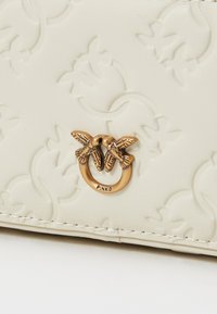 Crème faux leren portemonnee met reliëfpatroon, voorzien van een goudkleurige sluiting met twee vogels en "PINKO" gegraveerd. Textuurafwerking.