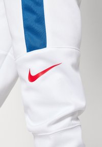 Pantalons de sport blancs avec une bande texturée bleue et un logo Nike swoosh rouge sur la jambe inférieure. Tissu doux avec des poignets côtelés.