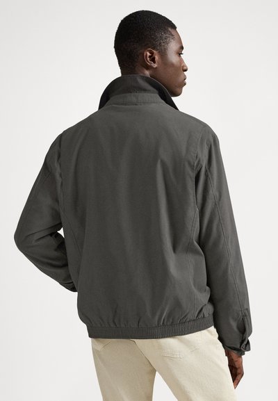 Chaqueta gris y ligera con dobladillo y cuello acanalados. Presenta una textura suave y un ajuste holgado, usada sobre pantalones de color claro.