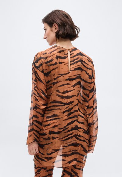 Γυναίκα με κοντά καστανά μαλλιά που κοιτάζει πλευρικά, φορώντας διάφανο καφέ και μαύρο μακρυμάνικο μπλουζάκι με ρίγες τίγρης και ταιριαστό παντελόνι.