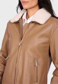 Bruine leren jas met een witte bontkraag, voorzien van een ritssluiting aan de voorkant en zijzakken. Soepele textuur en een aansluitend ontwerp.