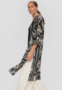 Schwarz-weißer Kimono-Cardigan mit floralem Muster, durchscheinendem Stoff, langen Ärmeln und offener Front; kombiniert mit weißen weit geschnittenen Hosen.