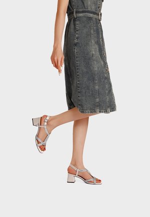 Donna che indossa un vestito midi in denim nero sbiadito con bottoni e sandali bianchi con cinturini, tacco largo aperto sulla punta, sollevando leggermente un piede.