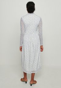 Robe à manches longues en tissu blanc transparent ornée de petits motifs bleus et jaunes, avec une taille cintrée et une jupe froncée.