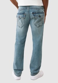 Jasnoniebieskie dżinsy o prostym kroju, z dwoma tylnymi kieszeniami z dekoracyjnym przeszyciem oraz z wyblakłym wykończeniem. Widoczne są białe sneakersy.