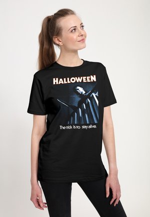 Młoda kobieta nosząca czarną koszulkę na Halloween z wizerunkiem zamaskowanej postaci trzymającej nóż z napisem „Sztuczka polega na przetrwaniu.”