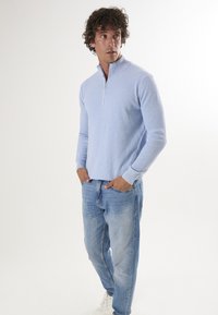 Pull à col zippé quart côtelé bleu clair, avec des manches longues, associé à un jean bleu clair et des baskets blanches. Look décontracté.