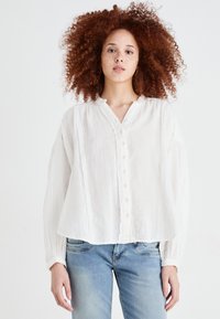 White, lightweight blouse with a button-down front, textured fabric, pleated details, and relaxed-fit sleeves, paired with blue jeans.