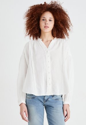 White, lightweight blouse with a button-down front, textured fabric, pleated details, and relaxed-fit sleeves, paired with blue jeans.