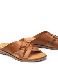 Sandalias deslizantes de cuero marrón con diseño de tiras trenzadas en cruz, suela plana y textura suave. Acabadas con un pequeño detalle metálico.