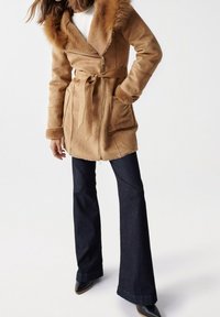 Tan faux pelsjakke med bred krave, lynlås og bælte. Kombineret med mørke, flared jeans og støvler med hæl. Glat tekstur.