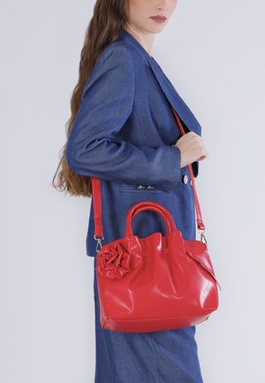 Kvinde iført blå blazer og nederdel holder rød læderhåndtaske med blomsterdetalje og skulderrem mod en ensfarvet baggrund.