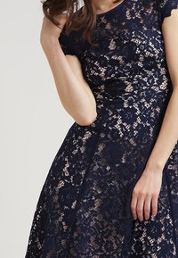 Námořnické modré krajkové šaty s jemnými květinovými vzory, krátkými rukávy, přiléhavým tělem a rozpínavou sukní s texturovaným povrchem.