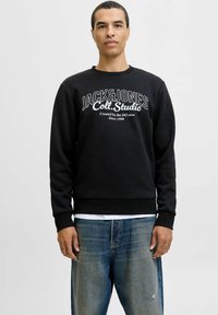 Czarna bluza z białym tekstem logo "JACK&JONES Coll. Studio" z przodu. Okrągły dekolt, ściągacze przy mankietach i u dołu, połączona z niebieskimi dżinsami.