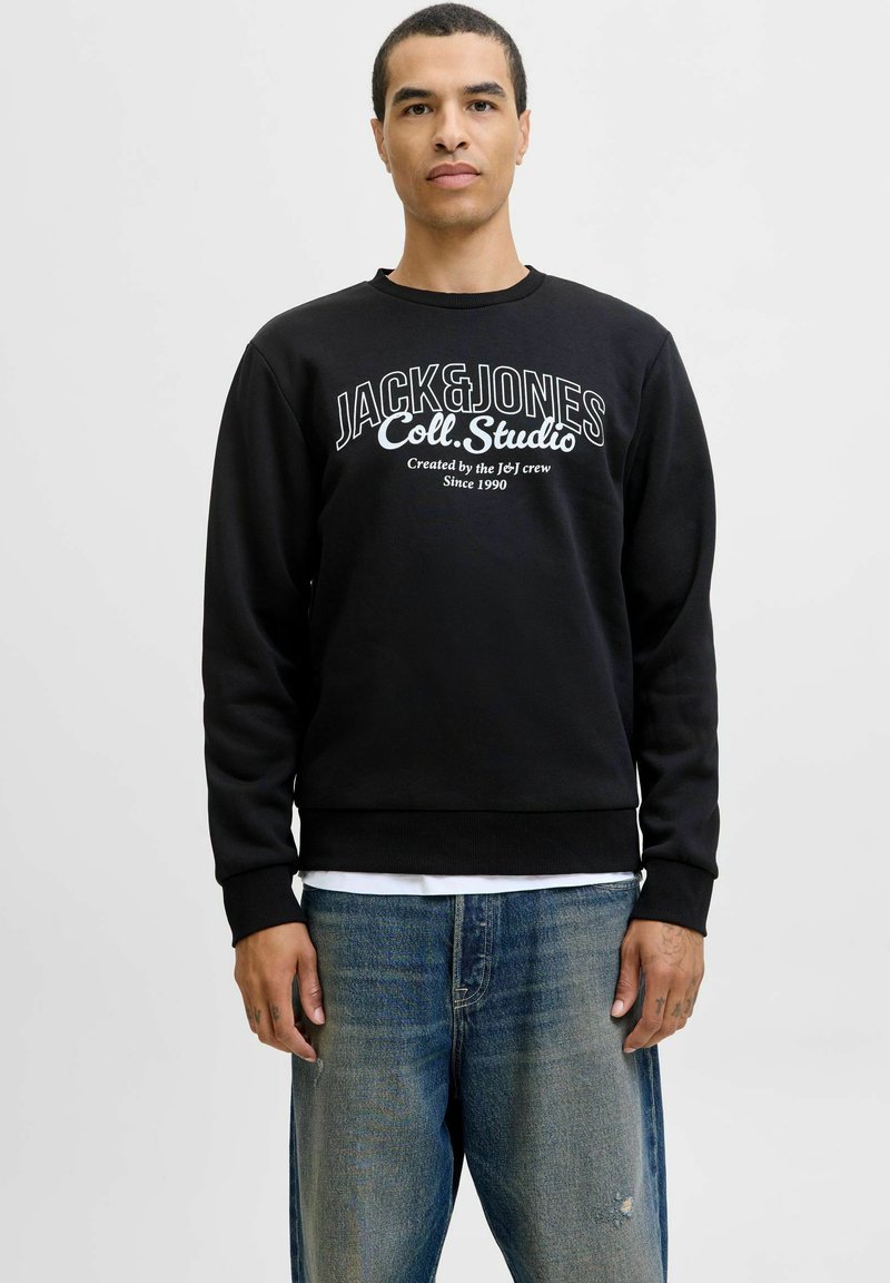 Czarna bluza z białym tekstem logo "JACK&JONES Coll. Studio" z przodu. Okrągły dekolt, ściągacze przy mankietach i u dołu, połączona z niebieskimi dżinsami.