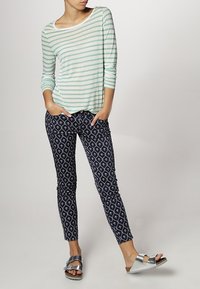 Světle zelený a bílý pruhovaný top s dlouhými rukávy, který je spojen s námořnickými kalhotami se vzorem geometrických tvarů, a stříbrnými plochými sandály.